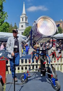 A man standing outdoors on a bright sunny day, operating a vintage carbon arc light with other exhibition stands in the background
