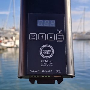 A black rectangular device with the logo for Power Gems and named GEMpsu plus user panel. In the background and out of focus are boats at anchor in a marina
